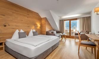 A modern hotel room with wooden paneling and large windows. It features a double bed, a seating area, and a desk.