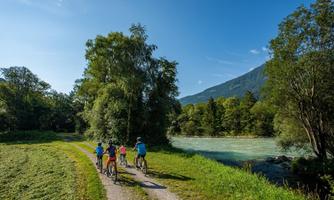 A group of cyclists is riding along a narrow path by the river. In the background, there are mountains and green trees visible.