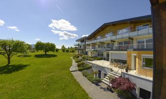 A modern building with balconies in a green environment. The sky is clear and sunny, surrounded by trees and lawns.