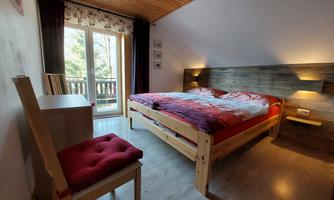 A cozy bedroom with a large bed and wooden details. Next to the bed is a table with a red chair and windows overlooking the outside.