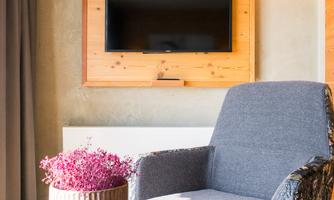 A cozy room with a gray armchair, a round table, and a television on the wall. A vase with colorful flowers is on the table.