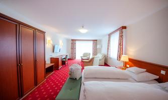 A spacious hotel room with two beds and a cozy seating area. The room has carpet flooring, large windows, and wooden cabinets.