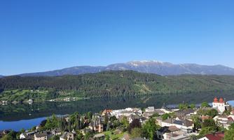 A picturesque landscape with a tranquil lake surrounded by mountains. In the foreground, small houses and plenty of greenery are visible.