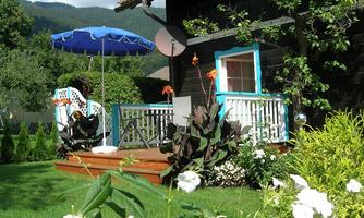 A beautiful garden with a wooden terrace and a blue sunshade. The surroundings are green and blooming, with a cozy atmosphere.