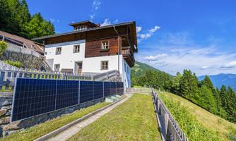 A modern house with a wood and plaster facade in a green setting. Solar panels are installed on the facade, and the sky is clear and blue.