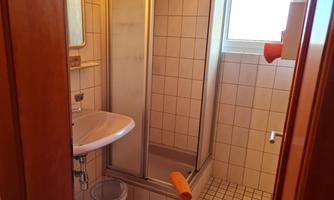 A simple bathroom with a shower, a sink, and a window. The walls are tiled and there is a small garbage bin in the room.
