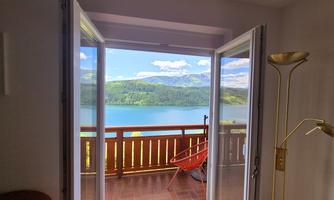 A beautiful view of a clear lake and green hills. The room has large windows with a red chair on the balcony.