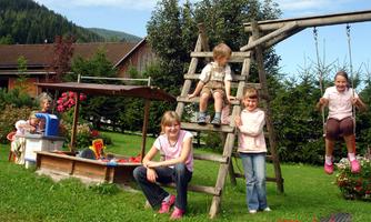 A group of children is playing outside. They are on a playground with a swing and a slide surrounded by trees and flowers.