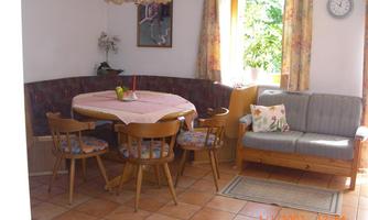 A bright dining area with a large bench and a round table. There are wooden chairs, a sofa, and large windows that let in plenty of light.