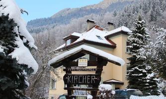 A cozy holiday home in winter, surrounded by snow and fir trees. In the background, snow-covered mountains and a clear sky can be seen.