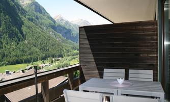 A cozy balcony with a table and chairs, surrounded by an impressive mountain landscape. In the background, tall mountains and a green valley are visible.