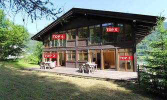 A modern wooden house with large windows and a terrace. The garden is green, surrounded by trees and mountains.