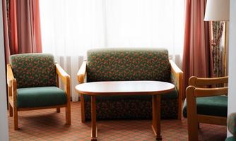 A cozy living area with two green chairs and a table. The windows let in plenty of daylight and the curtains are bright and friendly.