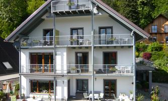 A modern house with several balconies and a terrace.  
Surrounded by green trees and flowers.