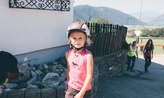 A little girl with a helmet is running outside in a rural area. In the background, more people and a picturesque landscape can be seen.