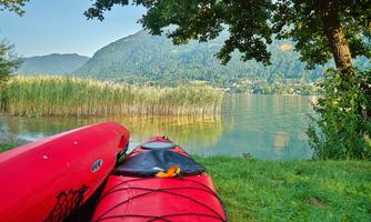 Two red kayaks are lying by the shore of a tranquil lake. In the background, mountains and green shoreline vegetation are visible.