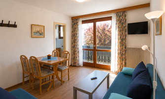 A bright living room with a dining area and a cozy seating corner. Large windows provide a view of the balcony and let in plenty of daylight.