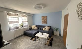 A modern living room with a large, dark sofa and a white coffee table. Bright curtains hang at the windows, and there is a decorative picture on the wall.