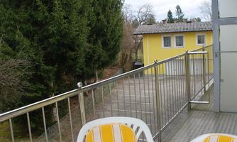 A balcony with two white chairs and a metal table. In the background, trees and a yellow building are visible.