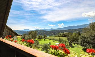 A picturesque landscape with green meadows and mountains in the background. Colorful flowers adorn the balcony and the sky is clear and blue.