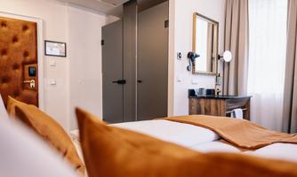 A modern hotel room with a comfortable bed and orange pillows. In the background, a mirror and a wardrobe are visible.