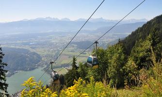 A cable car travels over green hills and a blue lake. In the background, mountains and a clear sky can be seen.