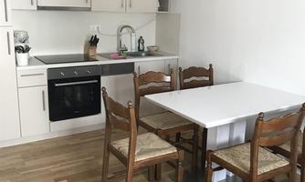 A modern kitchenette with white furniture and wooden flooring. A table with four chairs stands in the middle of the room.