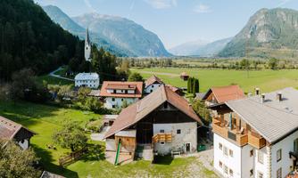 A picturesque landscape with traditional buildings and a church in the foreground. The surrounding mountains and the green field create a tranquil atmosphere.
