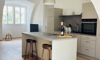 A modern kitchen with a beautiful kitchen island and stools. The light falls through large windows, creating a bright atmosphere.