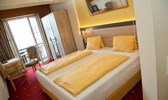 A cozy hotel room with two single beds and yellow cushions. There is a small table and a balcony that lets in plenty of light.