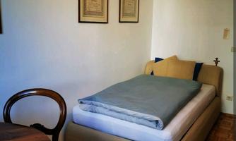 A simple bedroom with a bed and a small table. The walls are light and there are some pictures.