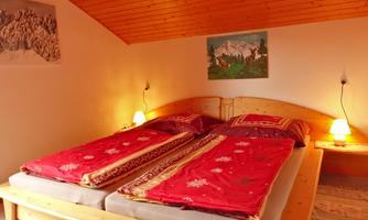 A cozy bedroom with a double bed and red bedding. The walls are decorated with pictures, and soft light illuminates the room.