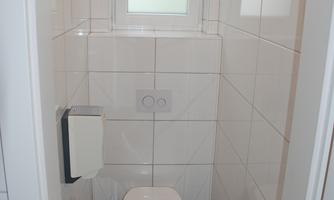 A modern bathroom with white tiles and a window. The toilet is simple and functional.