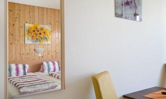 A cozy room with a bed and colorful cushions. In the background, there is a table with a chair and a wall with pictures.