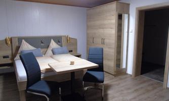 A modern bedroom with a double bed and a dining table. The room features wooden details and is well-lit.