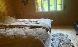 A cozy bedroom with wood paneling and two beds. A cut fur rug lies on the wooden floor and a window lets daylight in.