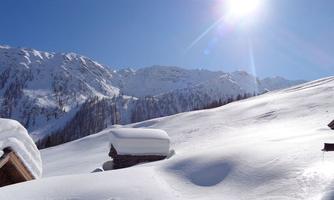A snow-covered landscape with mountains in the background and bright sunshine. In the foreground, there are small huts surrounded by smooth snow.