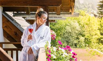 A woman in a bathrobe stands on the balcony holding a glass with a drink. In the background, green trees and colorful flowers can be seen.