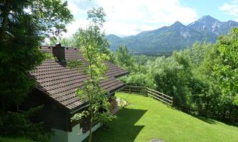 A beautiful house surrounded by trees and meadows. In the background, impressive mountains and a clear sky can be seen.