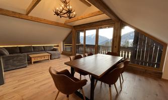 A cozy living space with a wooden ceiling, a large dining table, and a couch. Large windows offer a view of the mountains and the surrounding landscape.