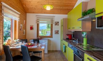 A modern kitchen with yellow cabinets and a dining table for four people. The room features wood paneling and large windows with a view of the greenery.