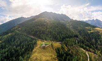 An impressive mountain landscape with dense forests and a green valley. In the foreground, a small building is visible, surrounded by nature.
