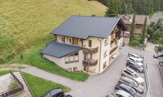 A two-story house in a quiet environment with a parking lot full of cars. In the background, green meadows and forests can be seen.