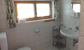 A bright bathroom with white tiles and windows with curtains. There is a sink, a mirror, and a toilet.