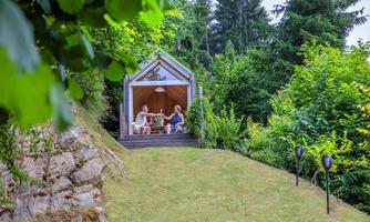 A cozy wooden structure stands in a green garden. Two people sit relaxed at it and enjoy nature.