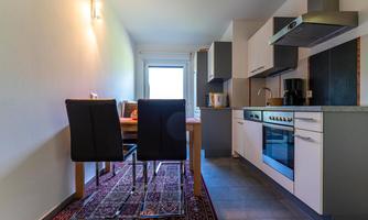 A modern kitchen with a dining table and chairs. There are bright walls, a stove, and windows that let in plenty of daylight.