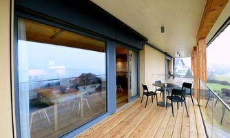 A modern balcony with a wooden floor and glass railing. There is a table with chairs and a view of the surroundings.