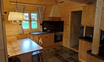 A cozy wooden kitchen with a dining area and a large window. The decor is warm and inviting, with wooden shelves and a modern stove.