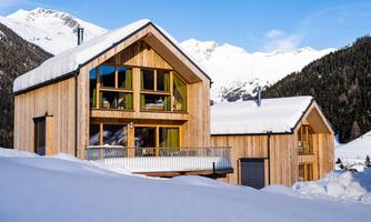 Two modern wooden houses in the snow, surrounded by mountains. The sky is clear and the landscape appears wintry and peaceful.