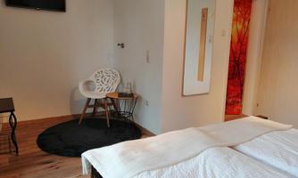 A modern bedroom with a double bed, a television, and a cozy armchair. The room features a warm wooden floor and an inviting atmosphere.